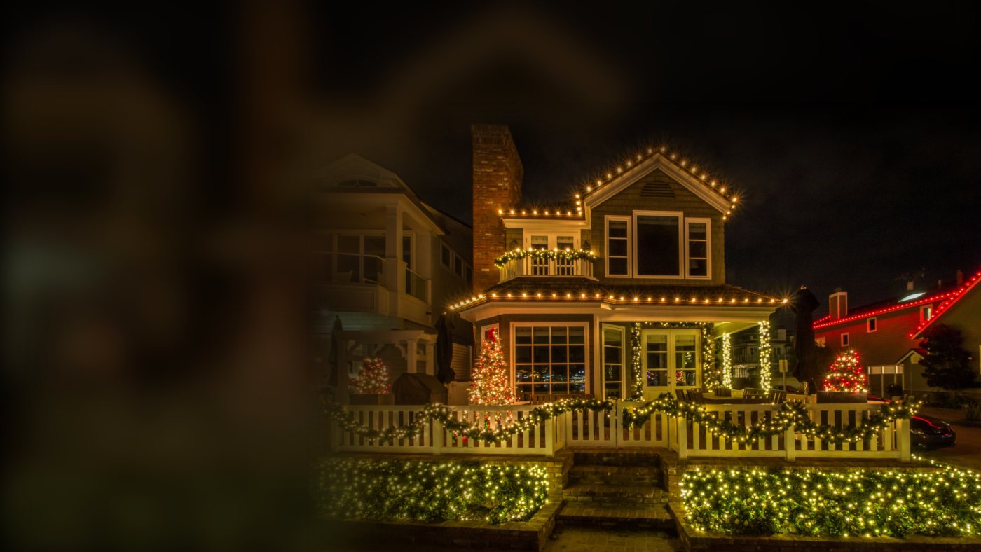 A two-story shingle home at night with warm white bulbs along every roofline, lit garland on the balcony and picket fence, glowing Christmas trees, wrapped porch columns, and net lights on the hedges