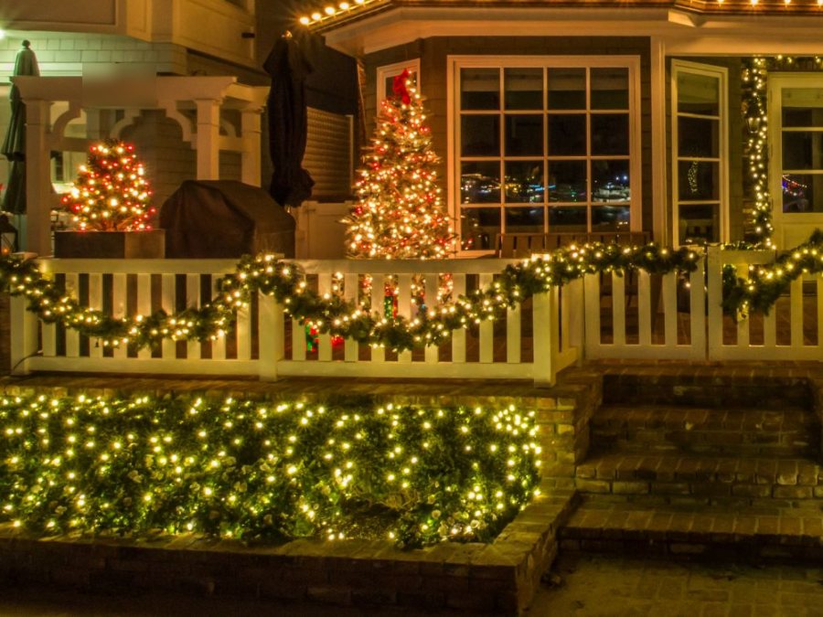 Net lights on the front hedges, a lit Christmas tree, and lit garland along a white picket fence